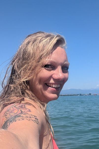 blonde Frau auf Wasser strahlt in Kamera, Tattoo auf Arm, Gardasee im Hintergrund