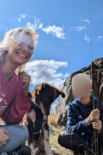 links blonde Frau, hockend, in der Mitte brauner Hund, rechts Junge hockend mit Stöcken in der Hand